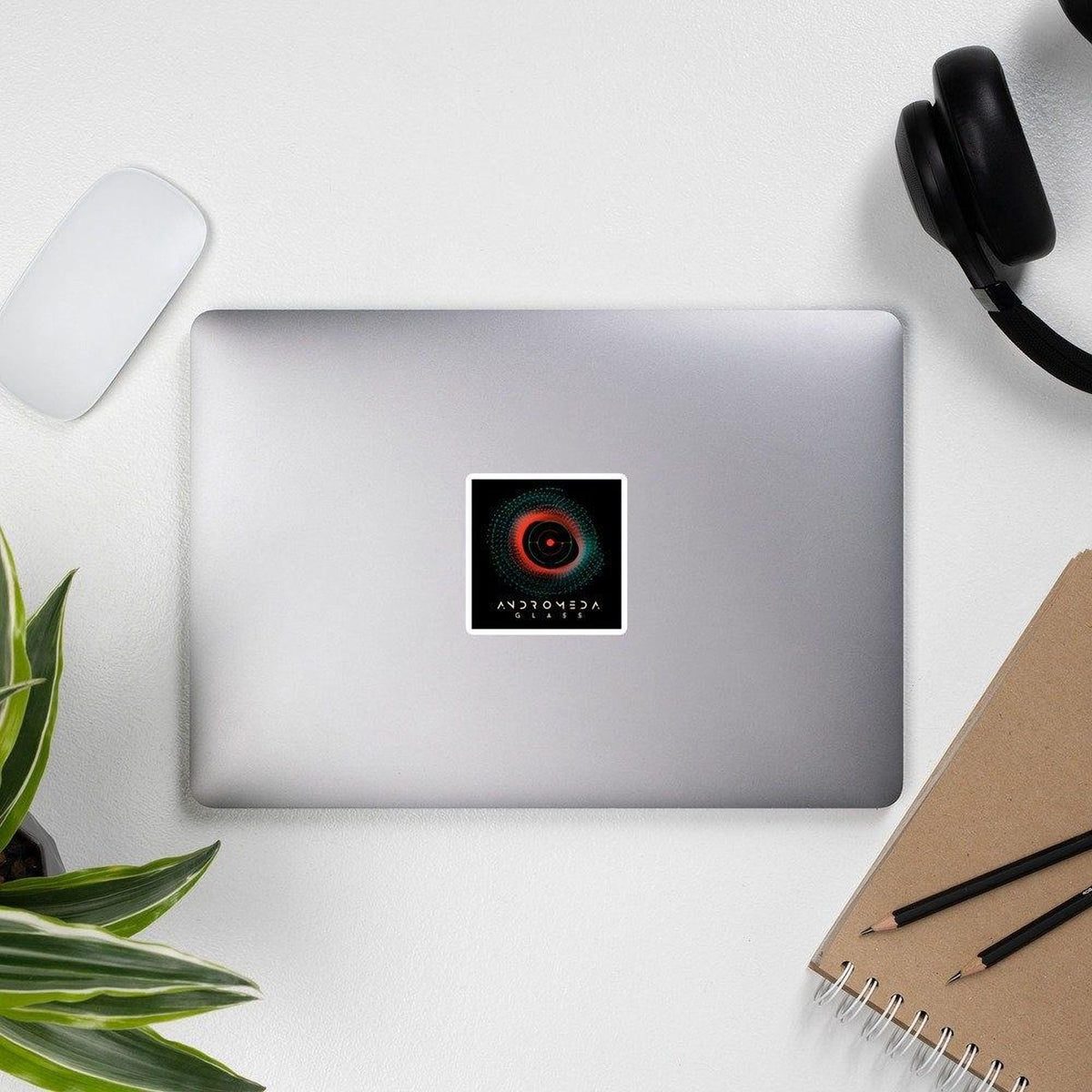 Silver laptop with a sticker on a white surface with a plant, notebook, and headphones.