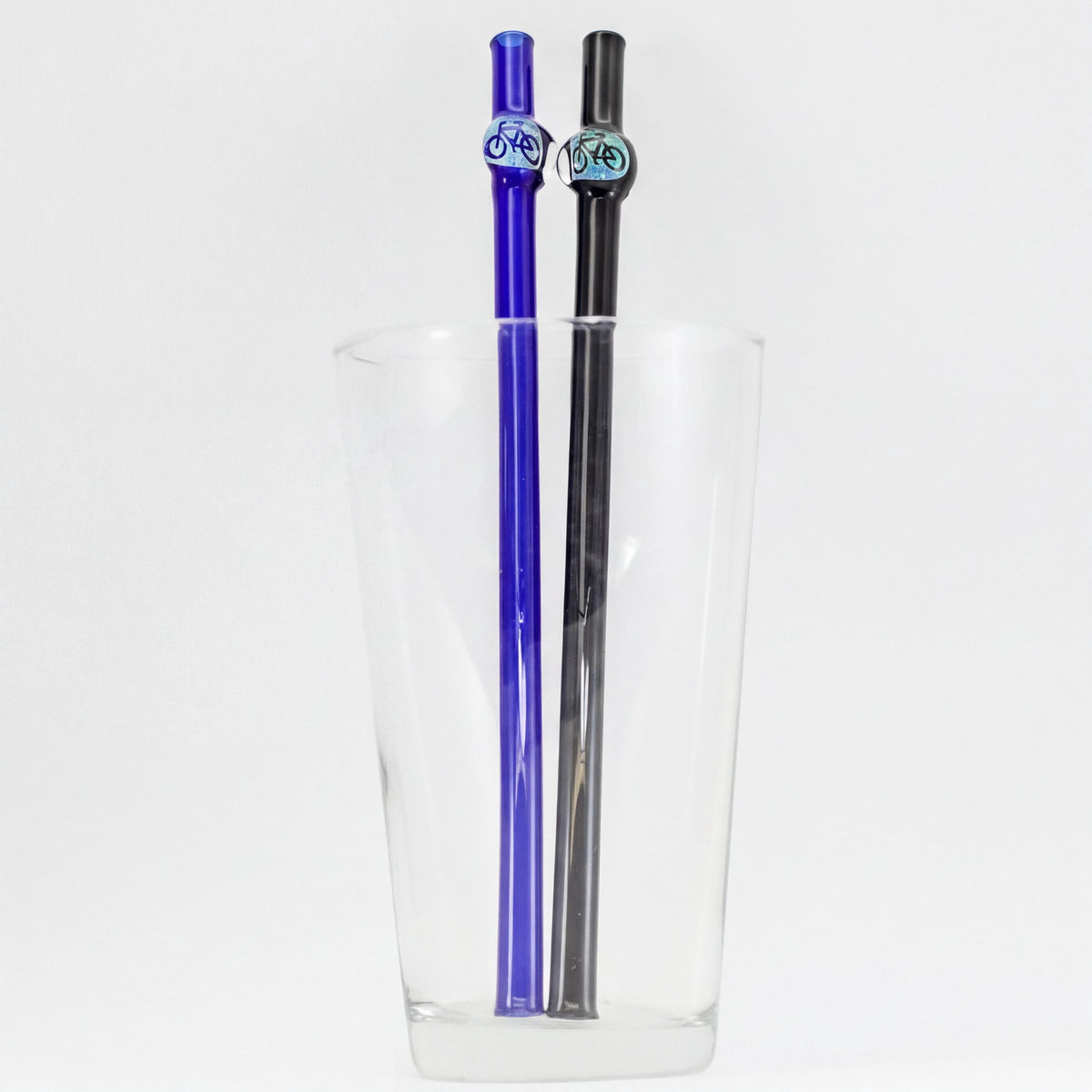 Two glass straws, one blue and one black, with decorative circular totem-like tops, displayed inside a clear glass cup on a white background.