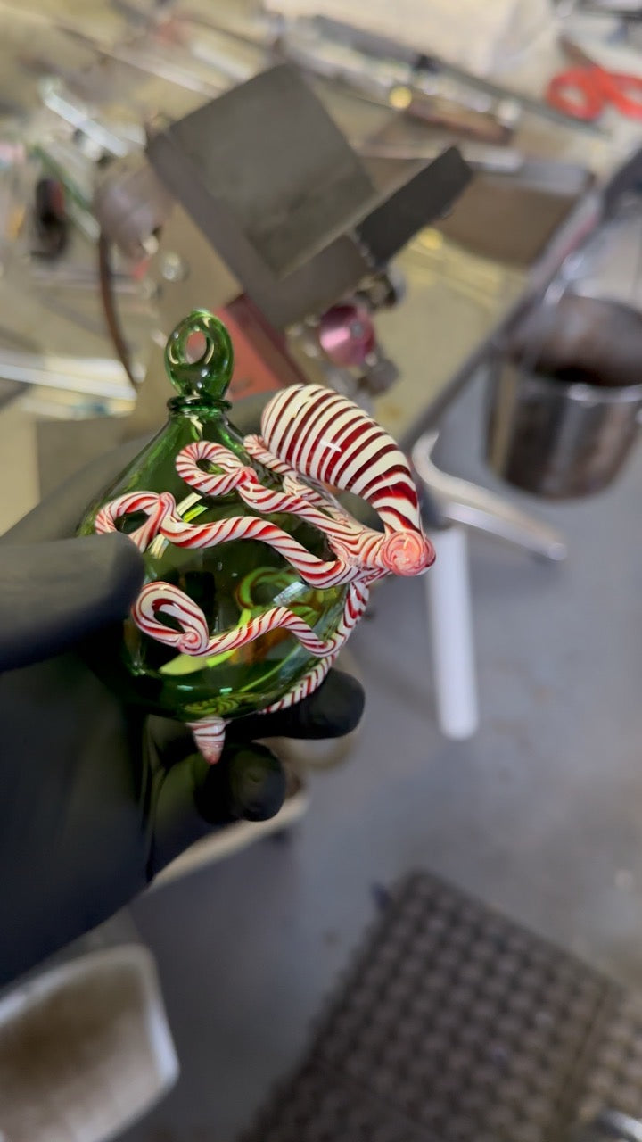 Black glove holding green globe ornament with red and white swirl octopus in a glass blowing studio. 