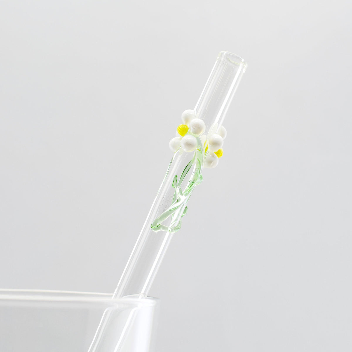 A clear glass straw with a small white daisy and yellow center decoration, shown resting against a light gray background.