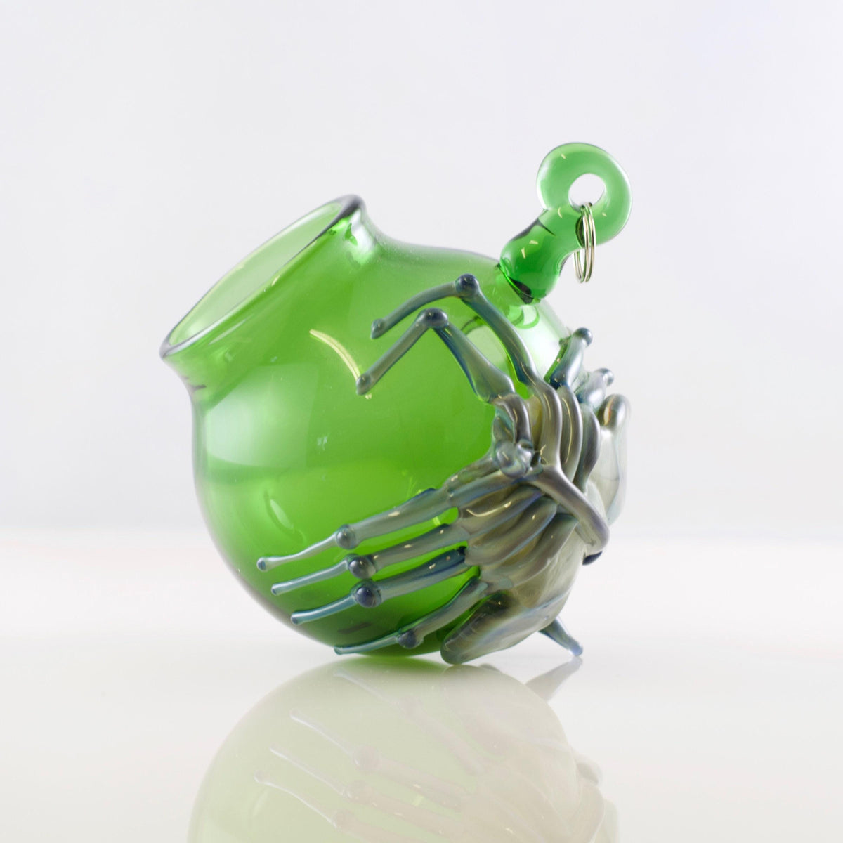 A green glass spider figurine wrapped around a matching green glass pitcher, with a small metal ring at the top of the pitcher, photographed on a white reflective surface against a white background.