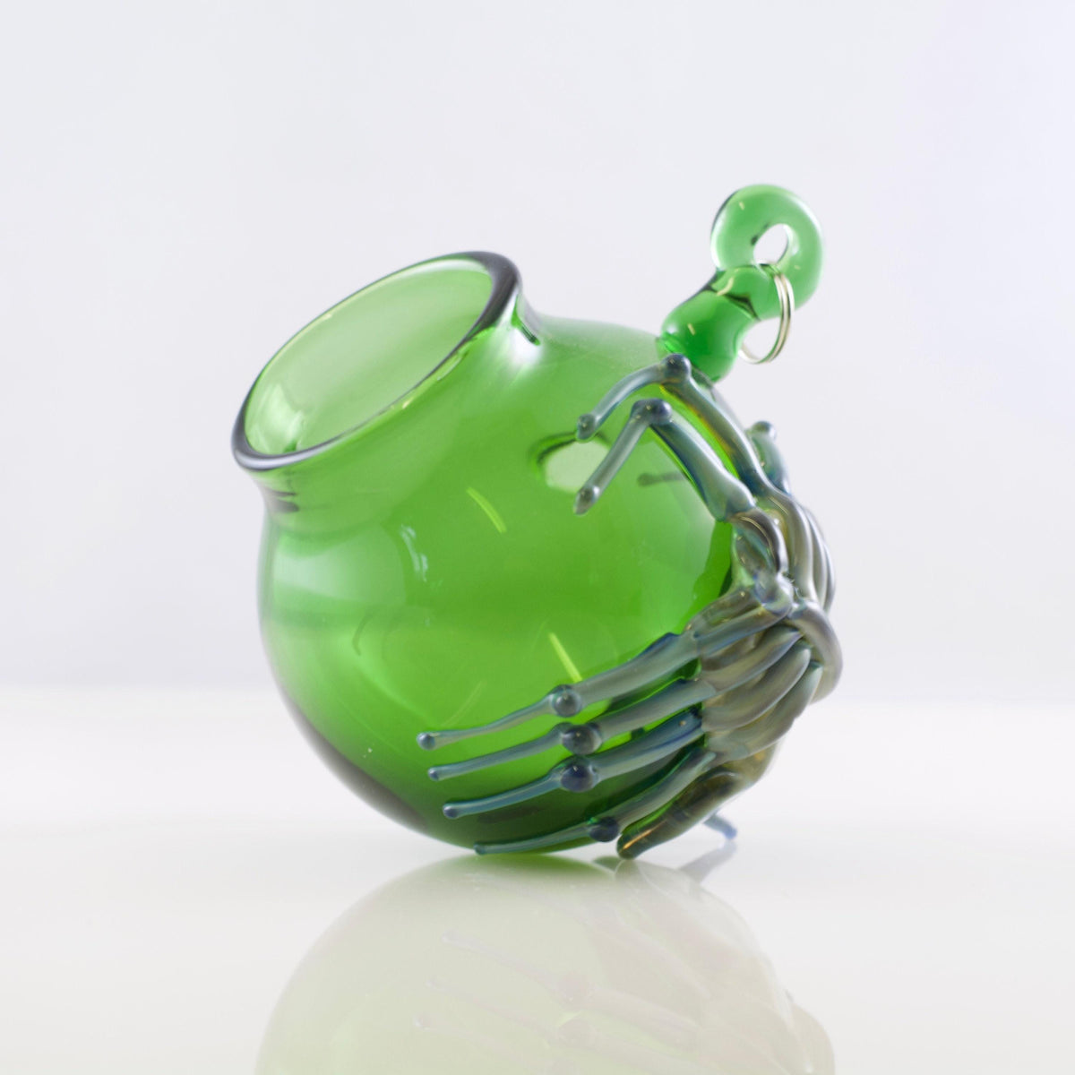 A green glass pitcher with a skeletal hand holding it, photographed on a clean white reflective surface against a white background.