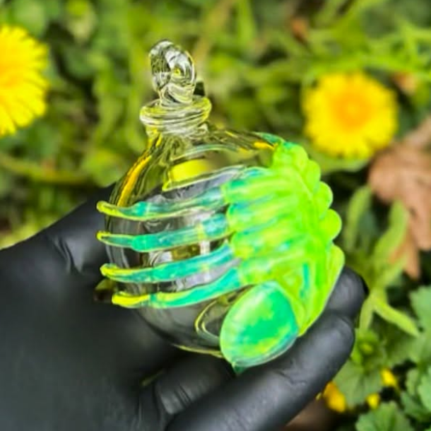 A hand wearing a black glove holds a small clear glass perfume bottle with a bright neon green and blue-green decorative design, set against a blurred background of green foliage and yellow flowers.