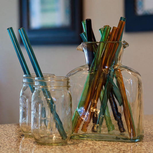 Three clear glass jars with matching glass straws in teal, green, brown, and black, arranged on a speckled beige countertop with a softly blurred indoor background.