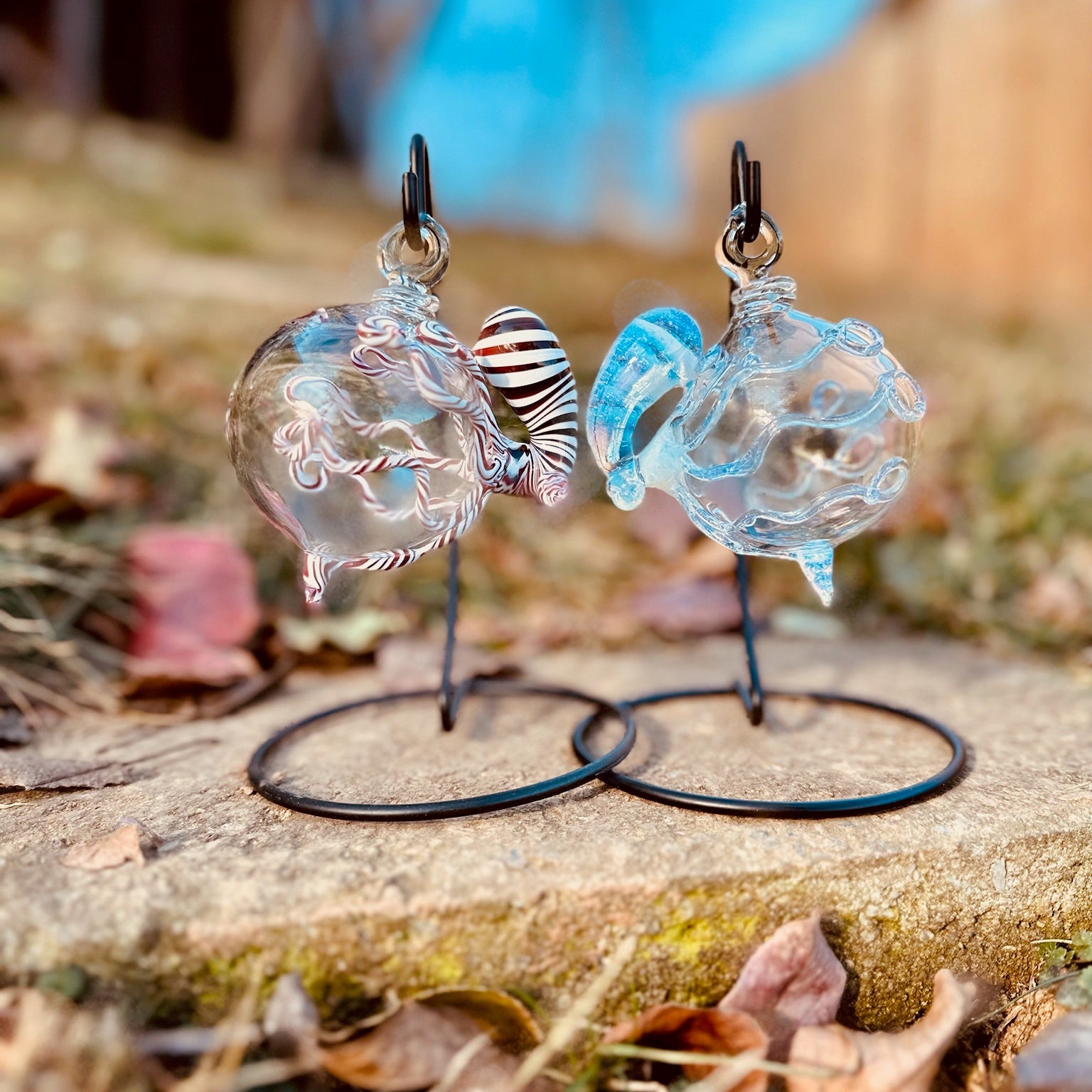 A pair of handmade glass dangle earrings displayed on black wire stands outdoors on a stone surface with dry leaves and a blurred natural background.