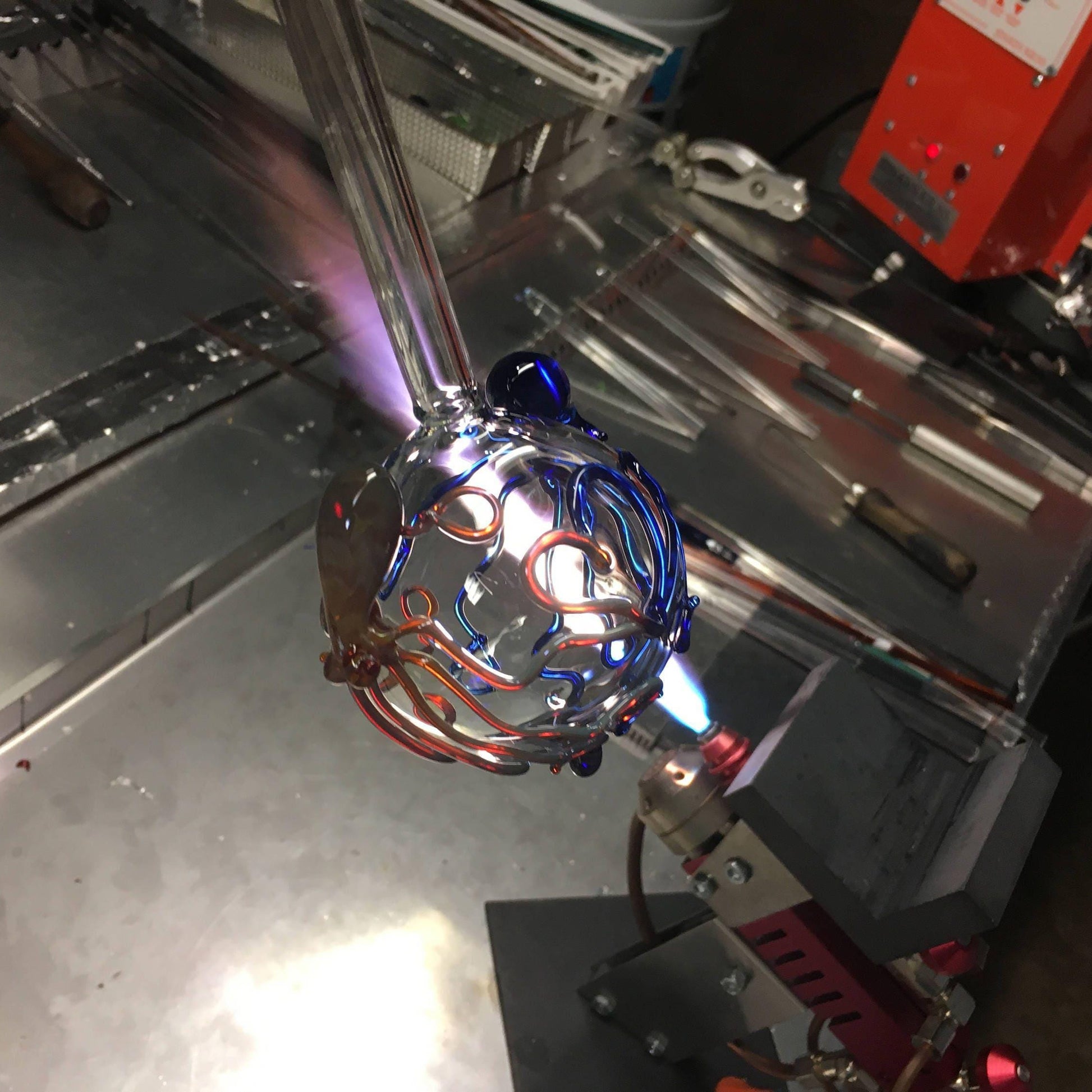 A close-up of a metallic decorative object with intertwined copper and blue wires, suspended near a bright torch flame in an industrial workshop setting.