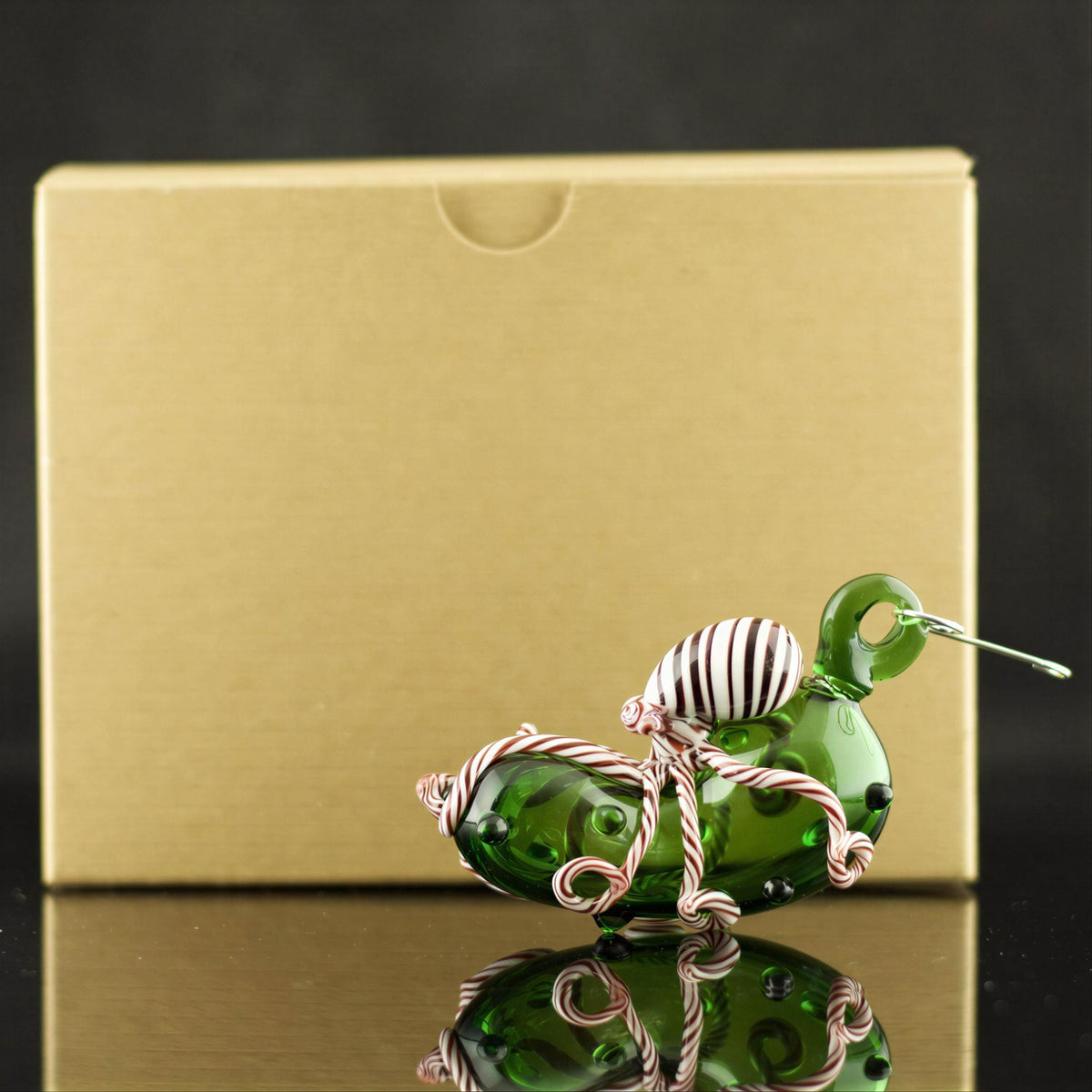 Green glass pickle ornament with candy cane swirl octopus on a reflective surface with a gold box in the background.