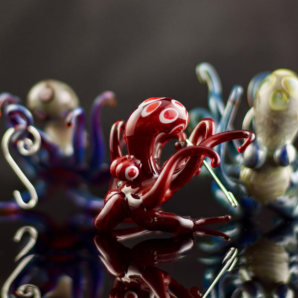 A glossy red glass octopus figurine with white circular accents, positioned centrally on a reflective dark surface with other colorful glass sea-creature figures blurred in the background.