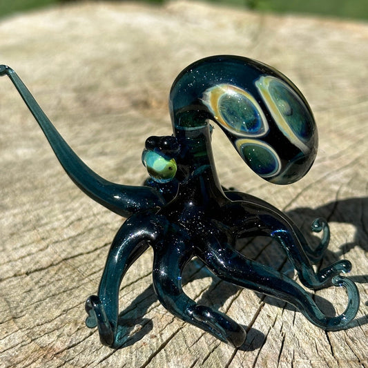A hand-blown glass octopus figurine with a glossy dark teal body and iridescent green, blue, and cream eye details, displayed on a weathered wooden surface outdoors.