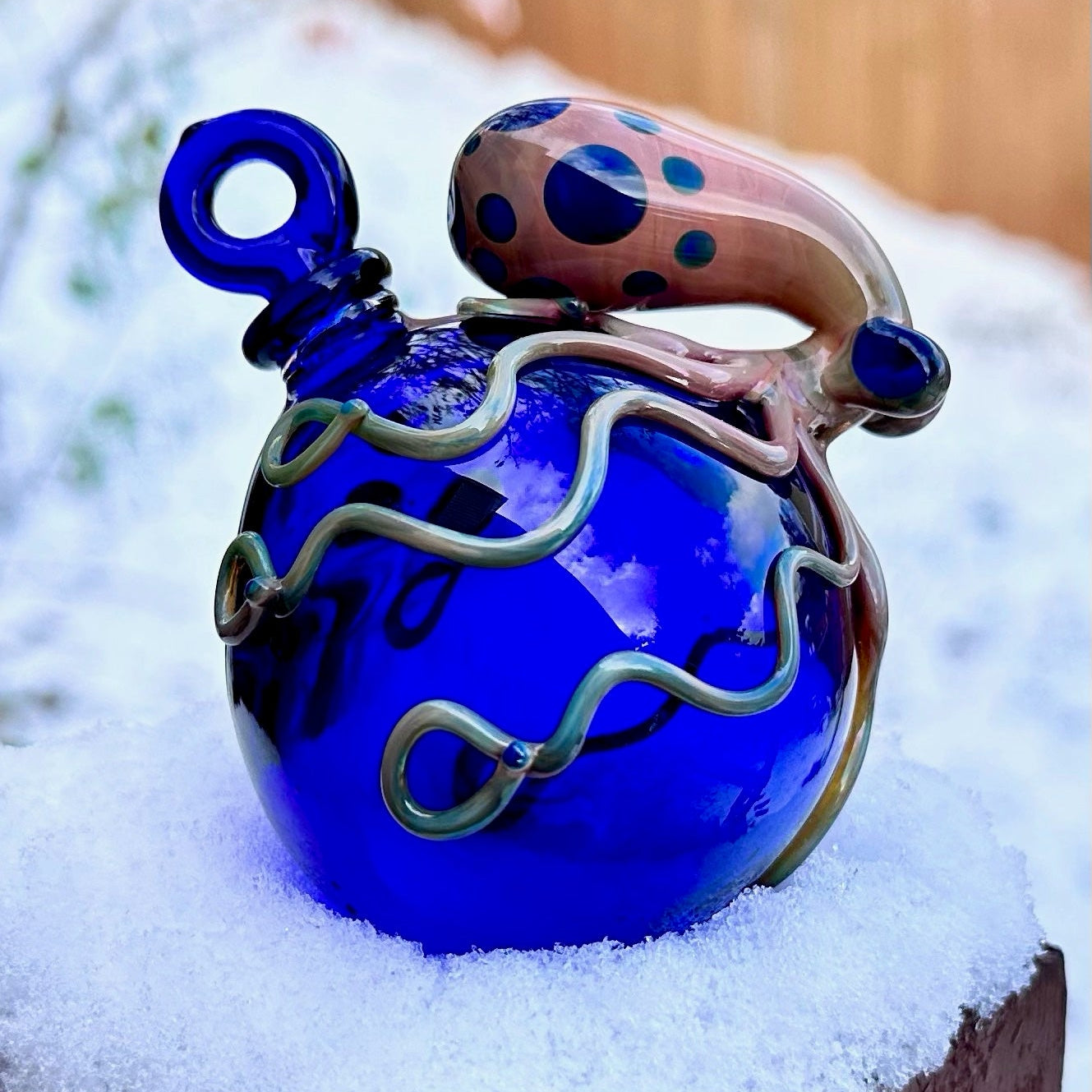 A cobalt blue glass ornament or decorative vessel with a loop handle and a curved handle decorated with dark blue polka dots, resting on a snowy surface outdoors.