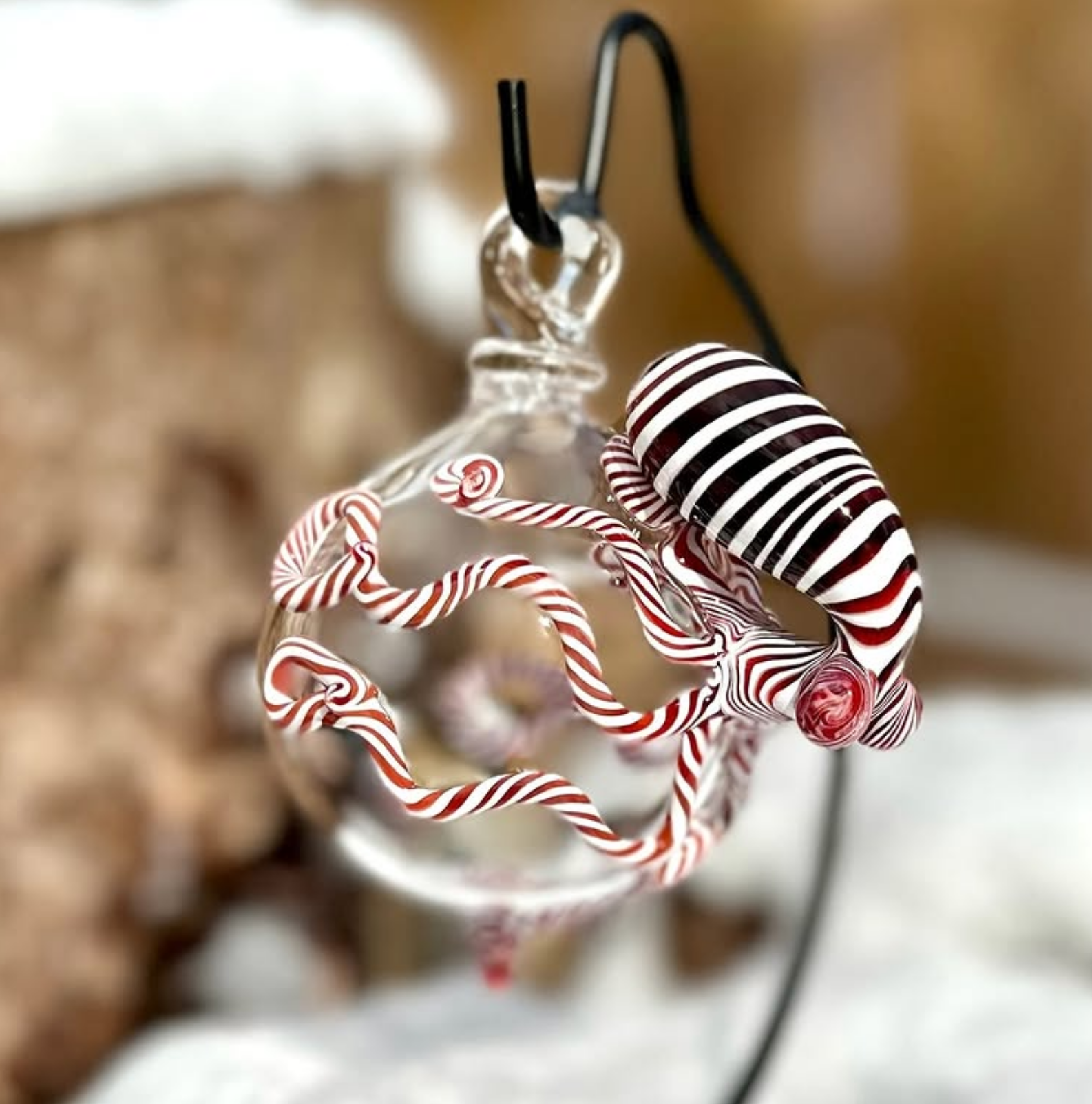 A clear glass ornament shaped like a Christmas tree branch with red-and-white striped candy canes and a small red bead, hanging against a blurred warm brown and white background.