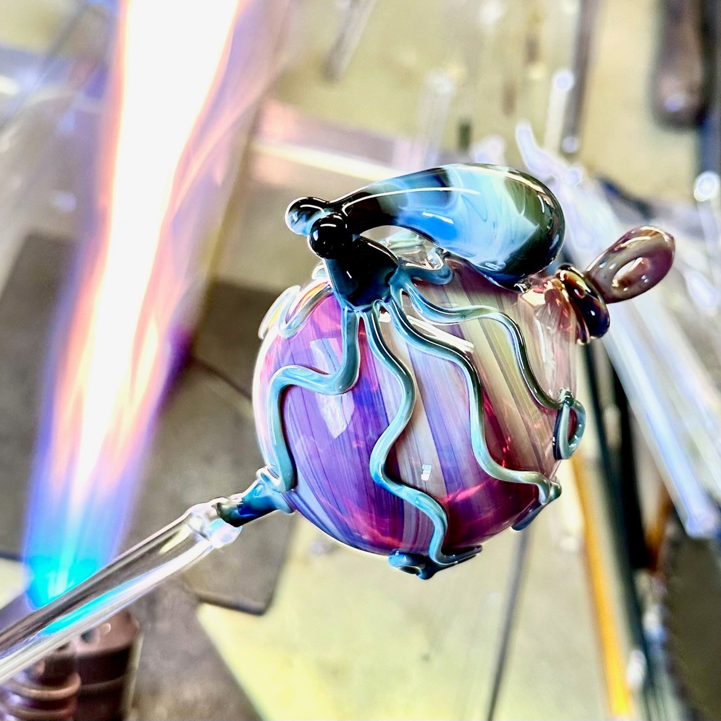 A hand-blown glass ornament with a purple and blue body, held on a glass rod under a blue flame in a glassblowing workshop.