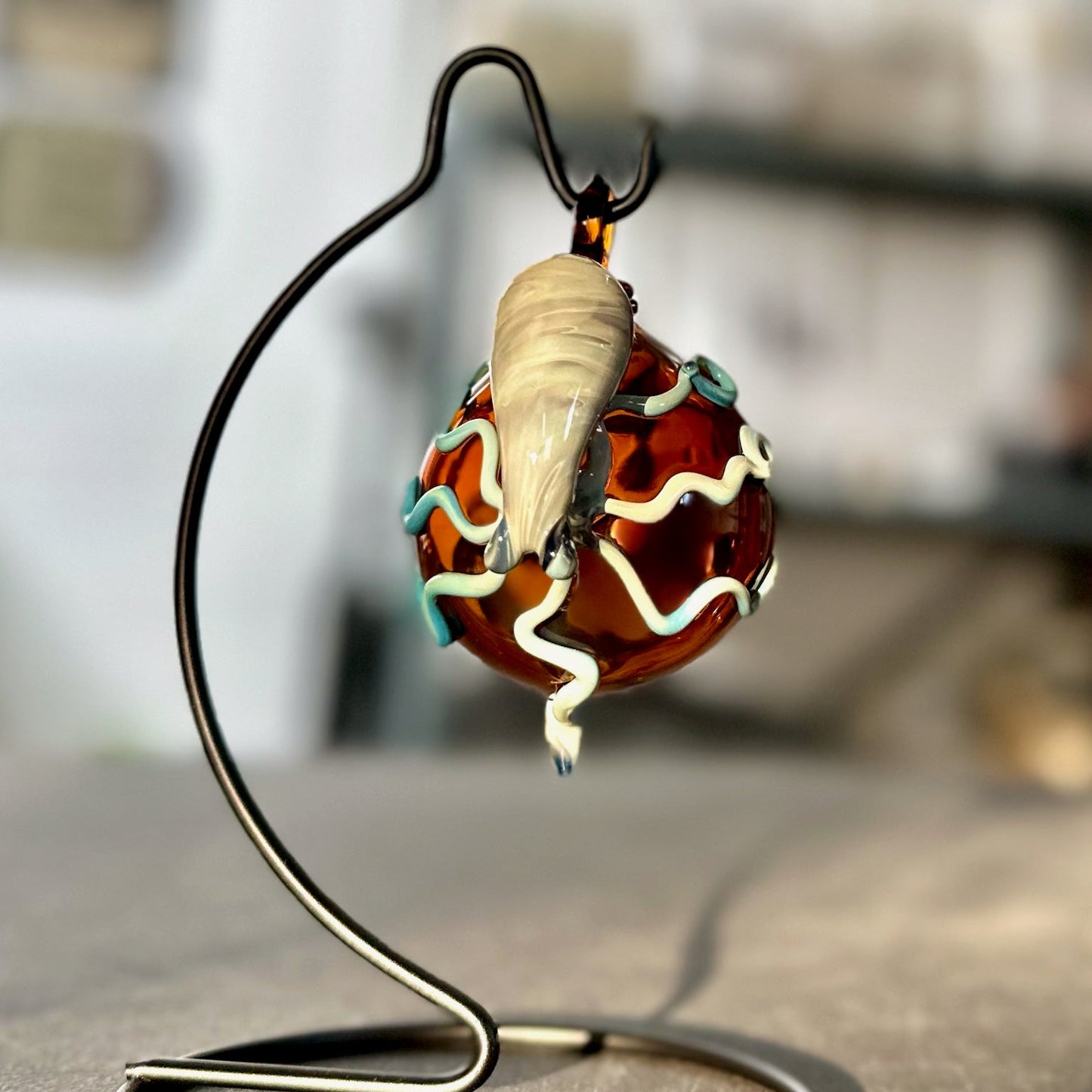 A handcrafted glass pendant with a translucent amber-brown body, decorated with white wavy lines, light blue accents, and a cream-colored shell, displayed on a black metal stand.