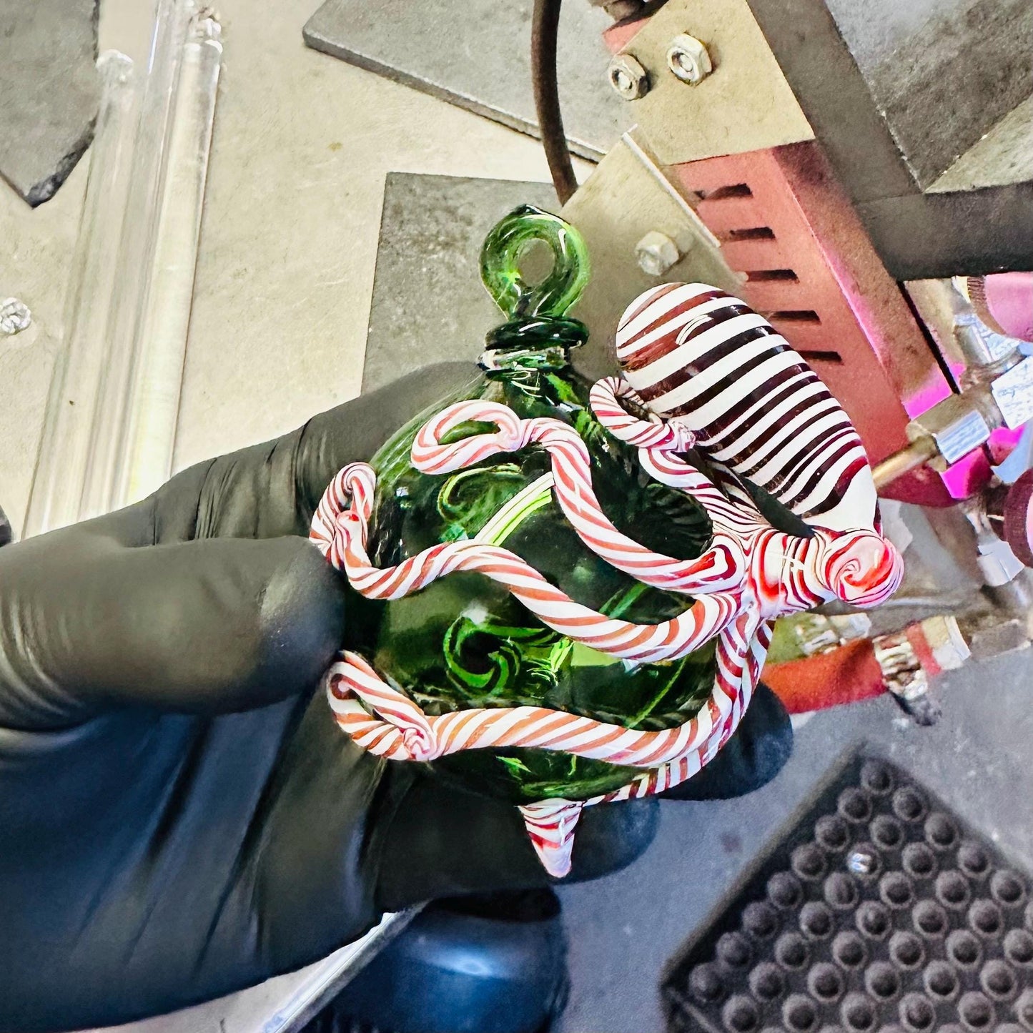 Hand holding a green glass ornament with candy cane design, wearing a black glove.