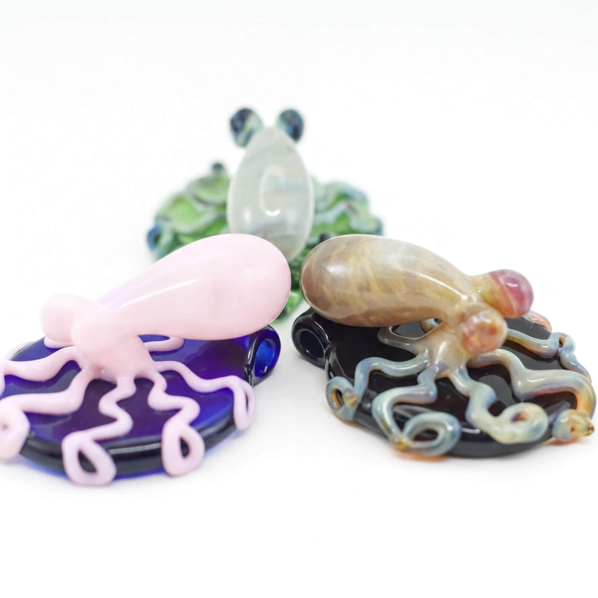 Three hand-blown glass octopus figurines on dark bases, displayed against a white background. The front-left octopus is light pink on a deep purple base, the front-right octopus is tan and gray on a black base, and a green octopus is visible in the blurred background.