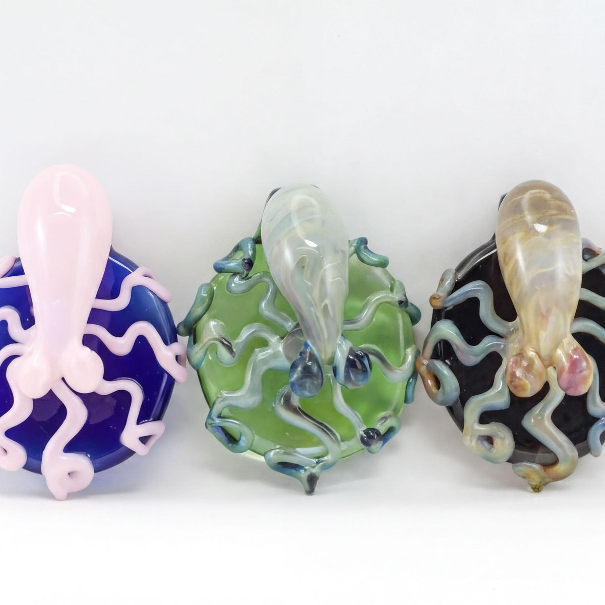 Three hand-blown glass decorative sculptures shaped like octopus-like creatures, displayed side by side on a white background. The left one is pink and blue, the middle one is green and blue, and the right one is black and beige with pink accents.