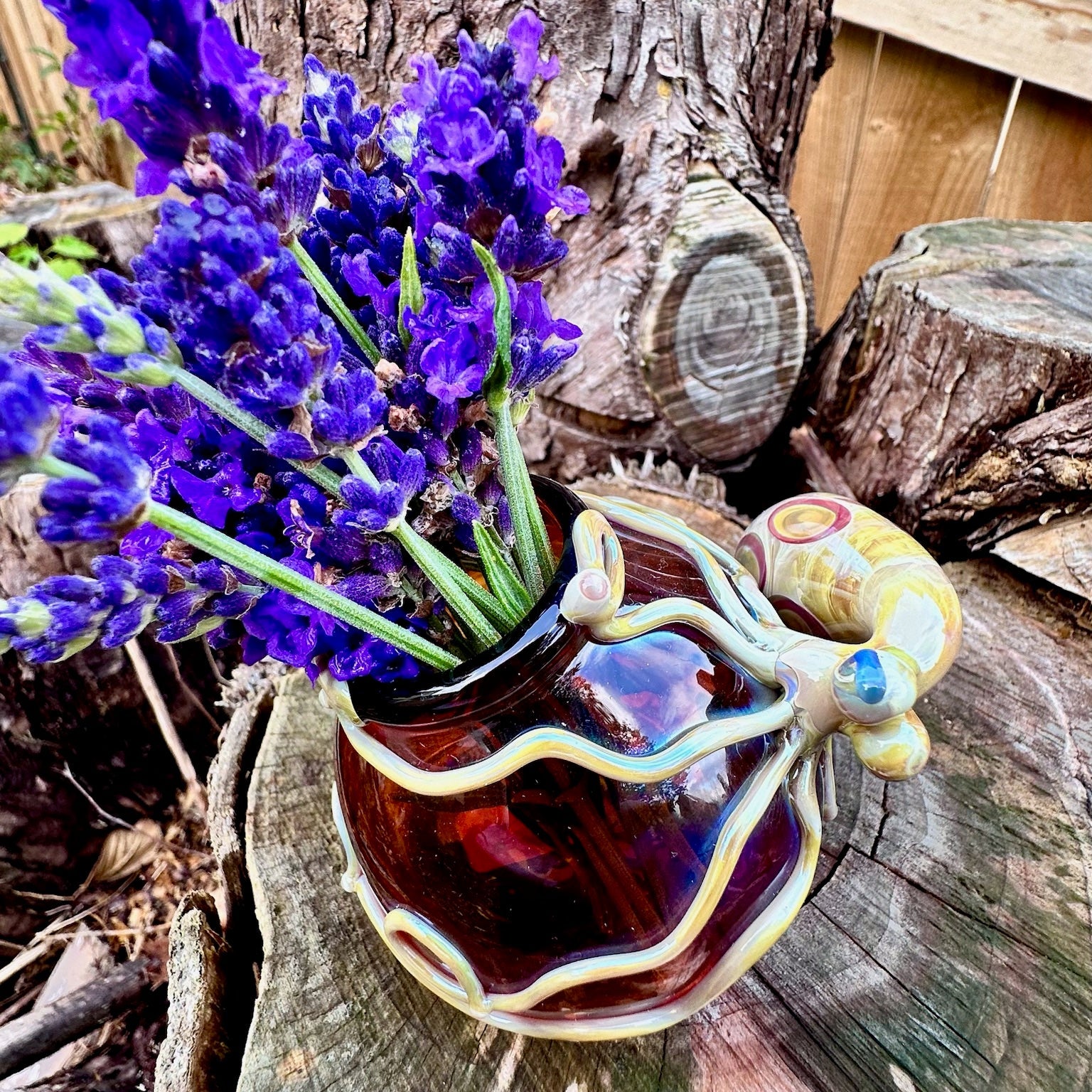 A small decorative ceramic vase with a bulb-shaped body, wavy rim, and a sculpted handle resembling a flower, filled with purple lavender stems, set on a rustic wood log outdoors.