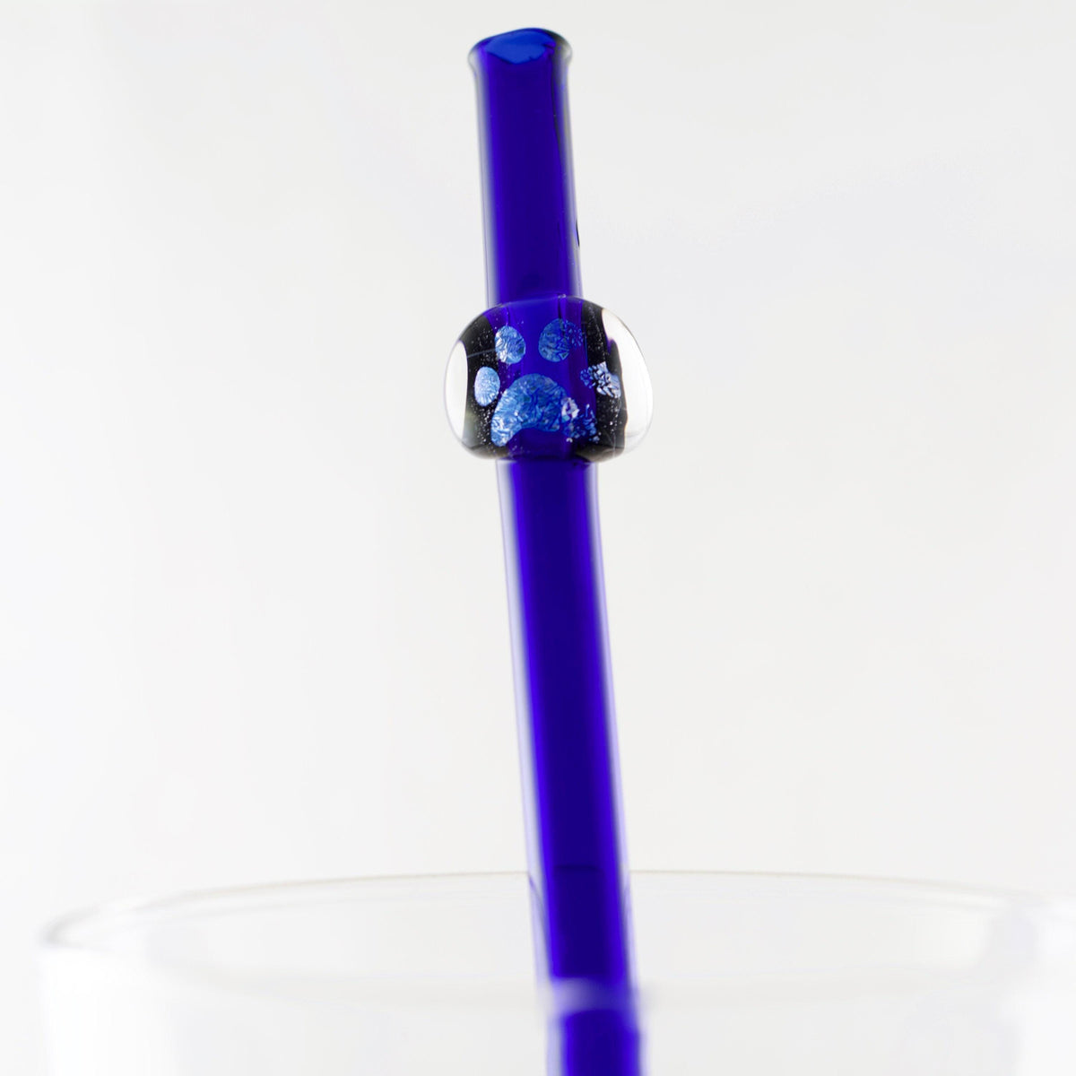 A close-up of a translucent cobalt blue glass straw with a clear bead featuring blue glitter paw prints, set against a white background.