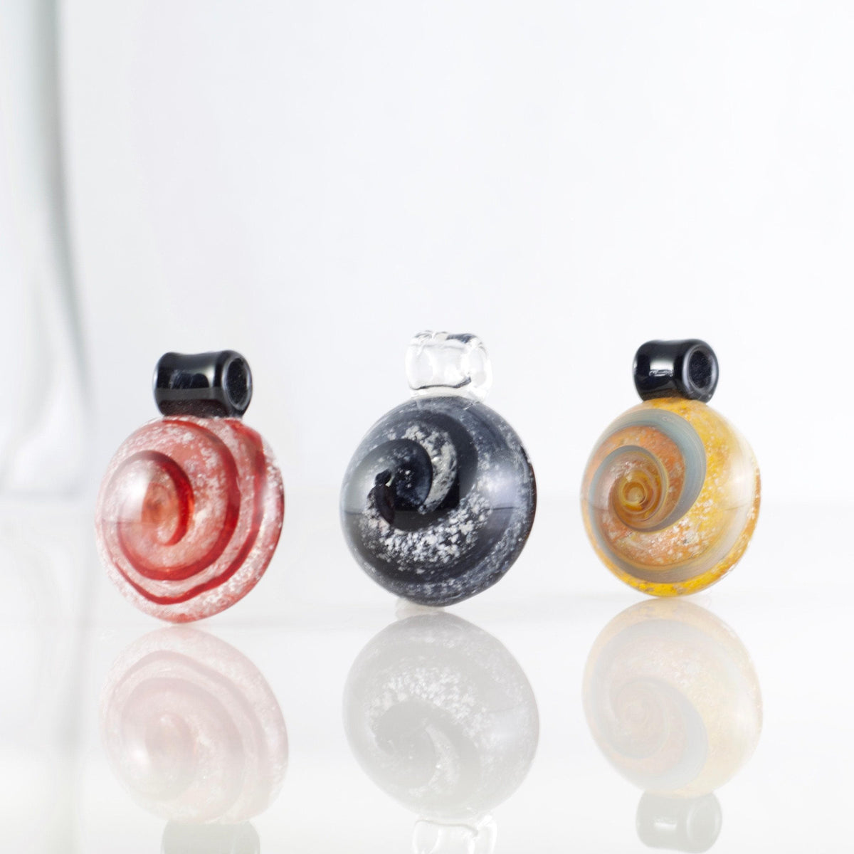 Three round glass bead charms with spiral patterns, displayed on a reflective white surface against a white background. The left charm is red and pink with white swirls, the center charm is dark gray and black with white glitter, and the right charm is yellow and orange with a gray spiral.