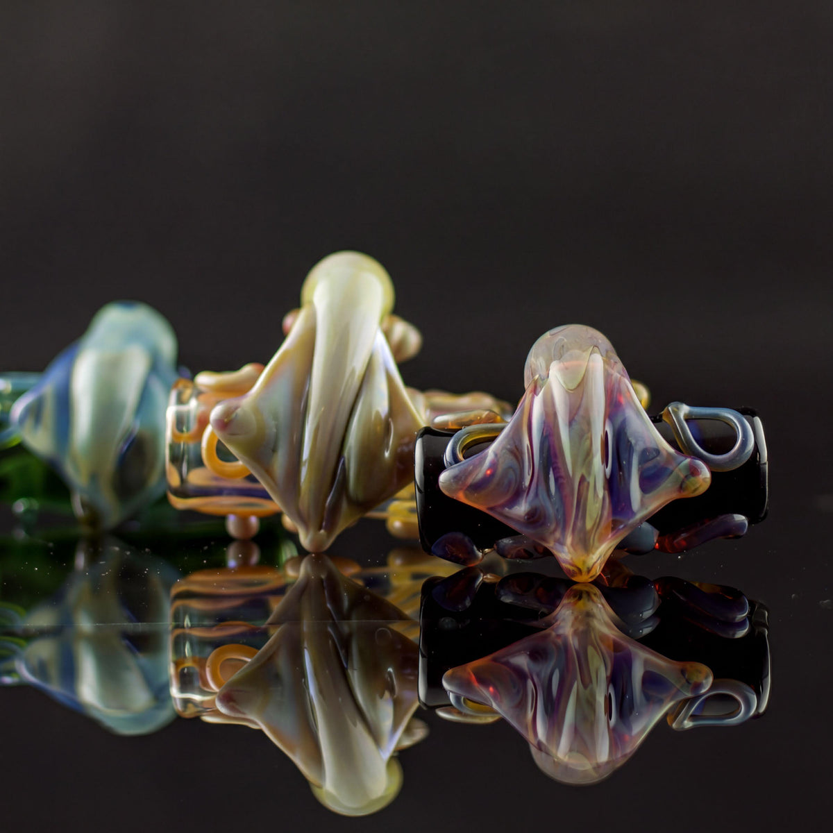 Three hand-blown glass lampwork beads with organic, spider-like forms, displayed on a reflective black surface against a dark background. The beads feature iridescent colors including cream, purple, blue, green, and black.