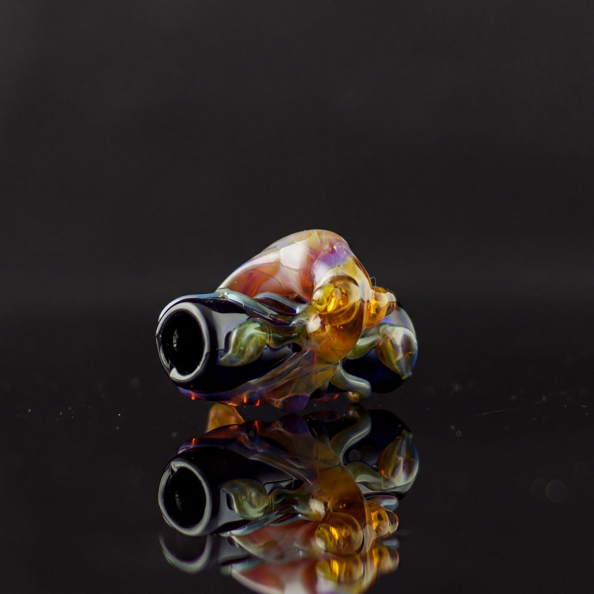 A hand-blown glass water pipe piece with a black mouthpiece and a bulbous body featuring swirls of amber, purple, green, and clear glass, resting on a reflective black surface.