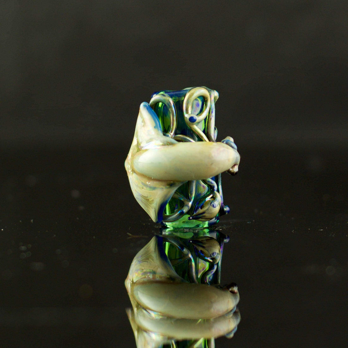 A hand-blown glass water pipe piece with a black mouthpiece and a bulbous body featuring swirls of amber, purple, green, and clear glass, resting on a reflective black surface.