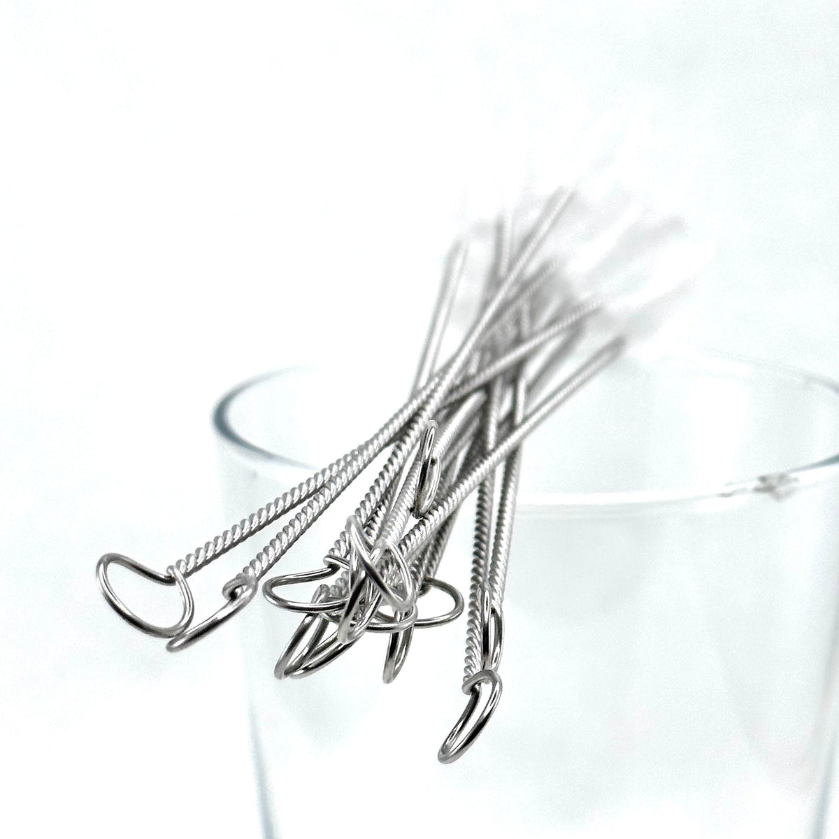 A set of stainless steel straw cleaning brushes with twisted shafts and looped ends, resting in a clear glass on a white background.
