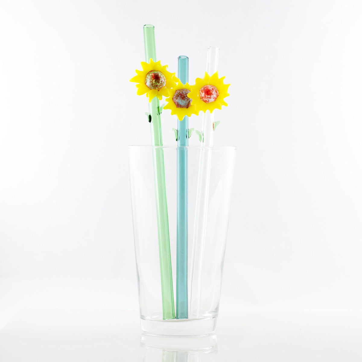 Three decorative drinking straws in pastel green, light blue, and white, each topped with a yellow sunflower, arranged inside a clear glass cup on a white background.
