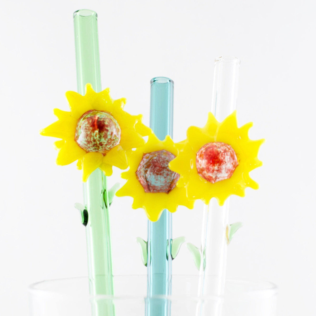Three decorative glass straws in mint green, light blue, and clear, each topped with a yellow sunflower-shaped ornament holding a small round candy, shown in a glass cup against a white background.