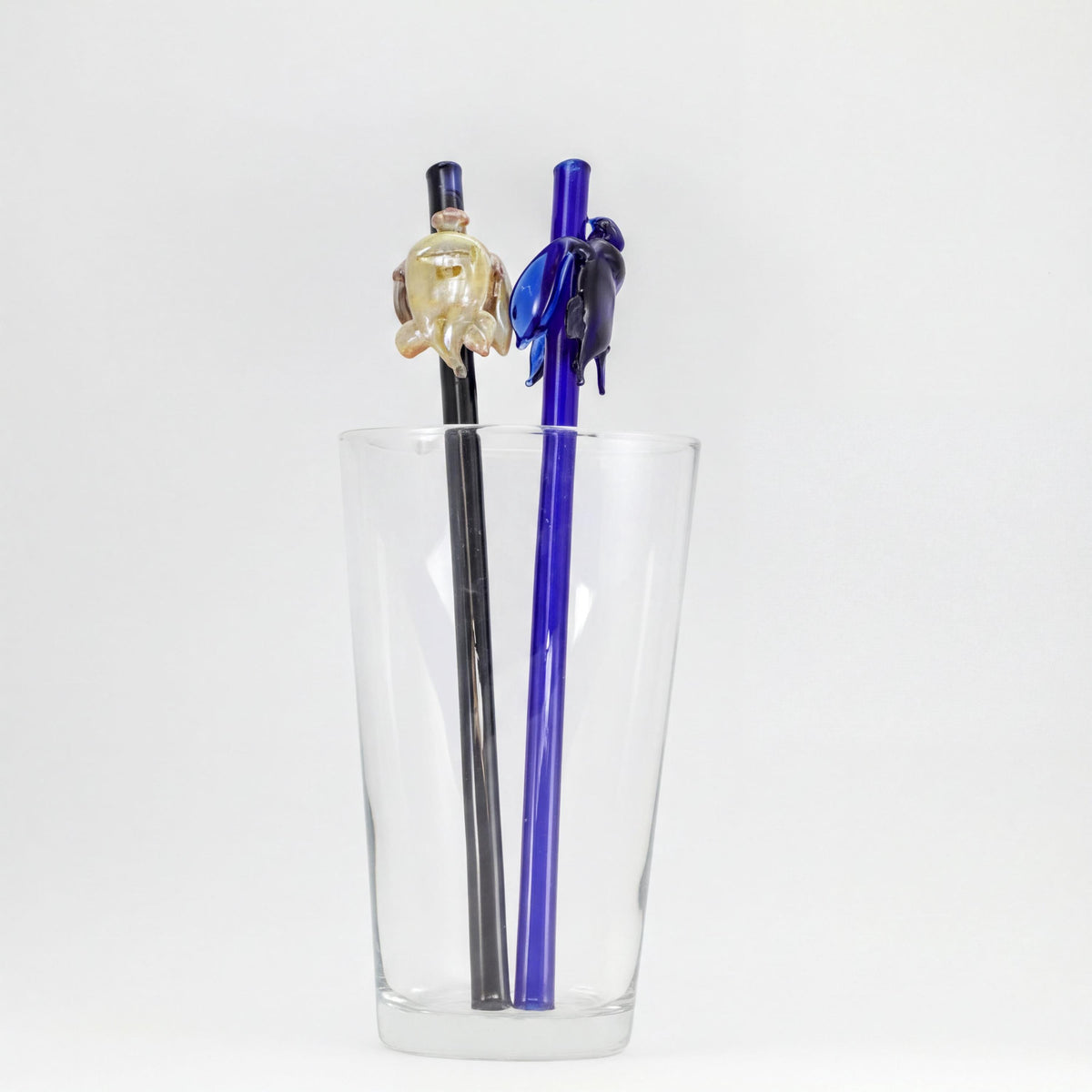 Two decorative glass straws with turtles one in black and one in blue, displayed inside a clear glass cup against a white background.